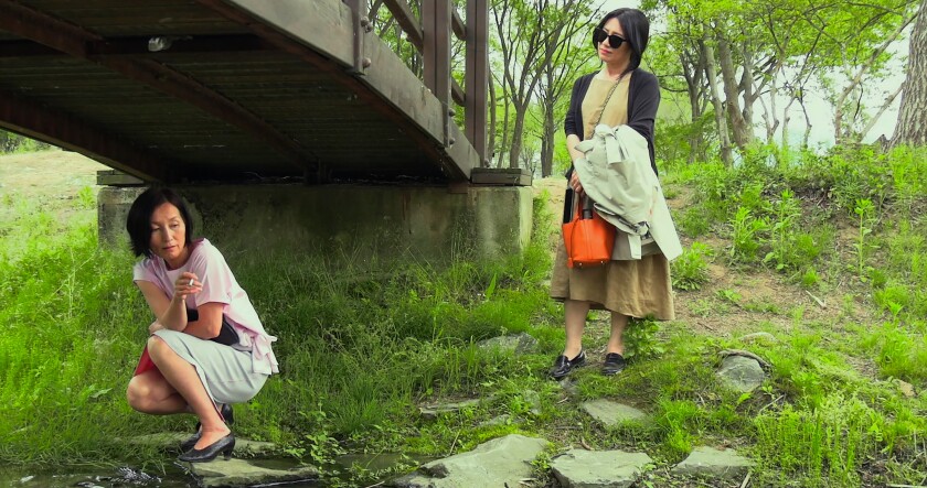 A woman crouches beneath a bridge over grass while another woman looks at her