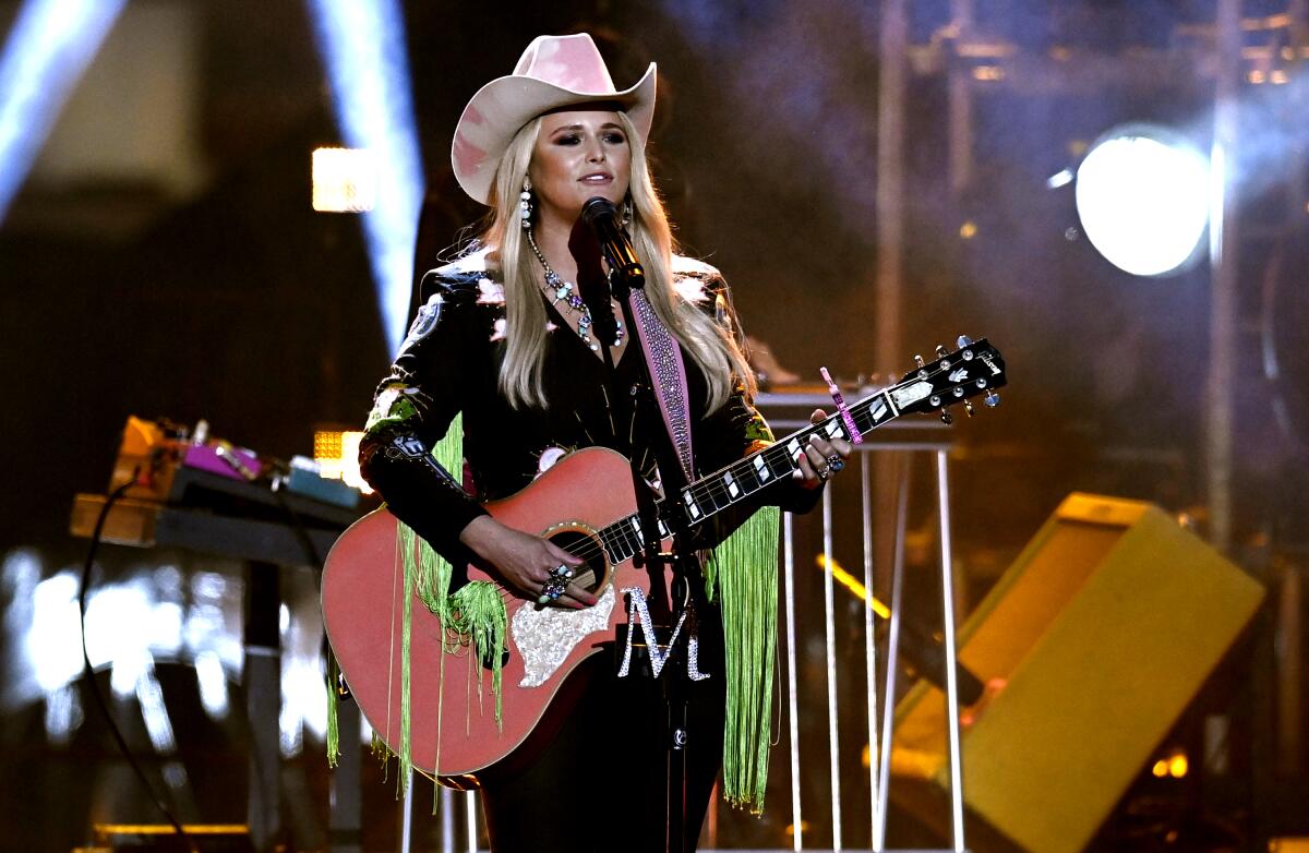A blond woman in a pink and white cowboy hat and black shirt with bright green fringe plays guitar onstage.