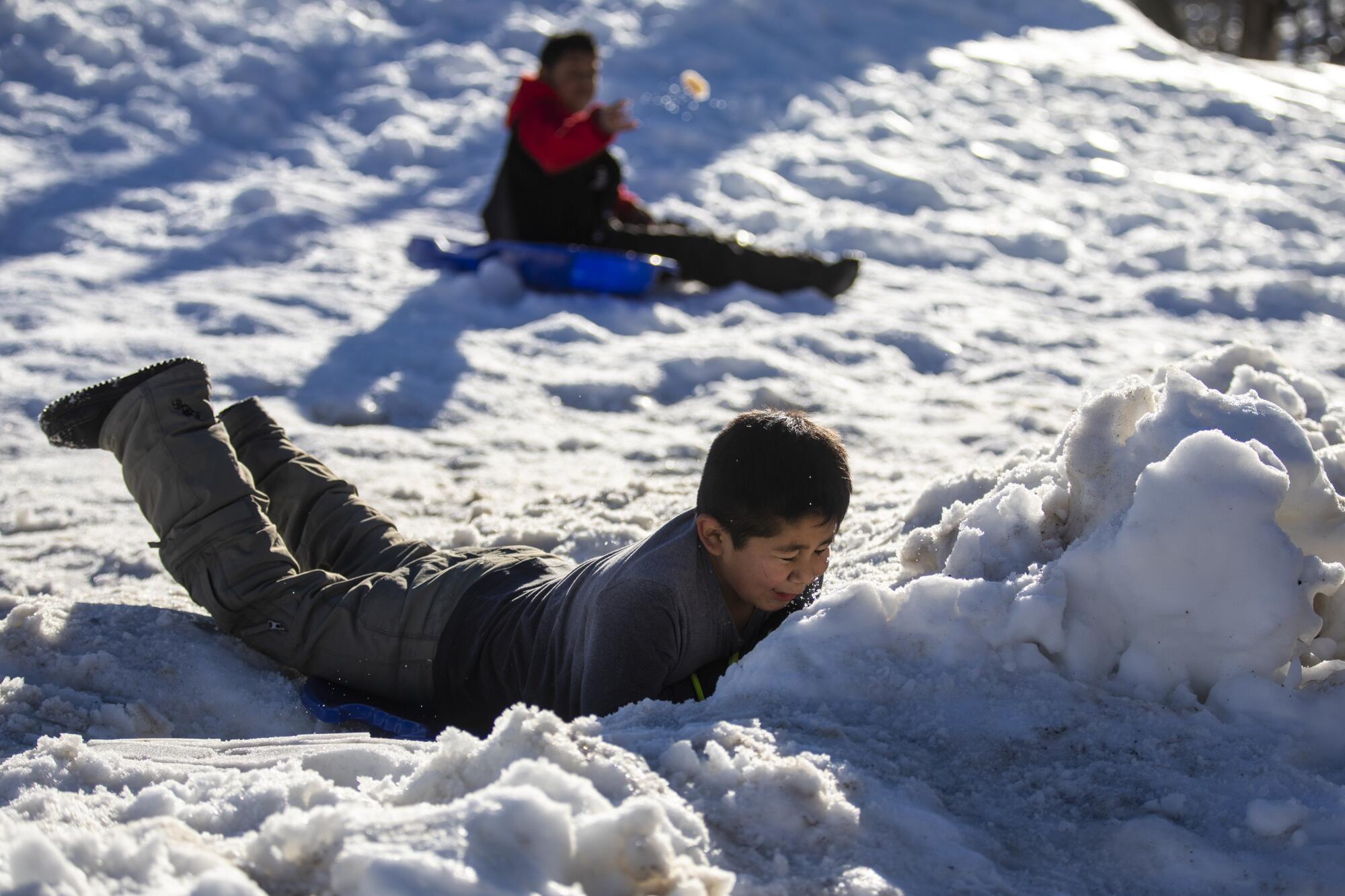 Two boys, one seated and another on his belly, ride sleds on packed snow.