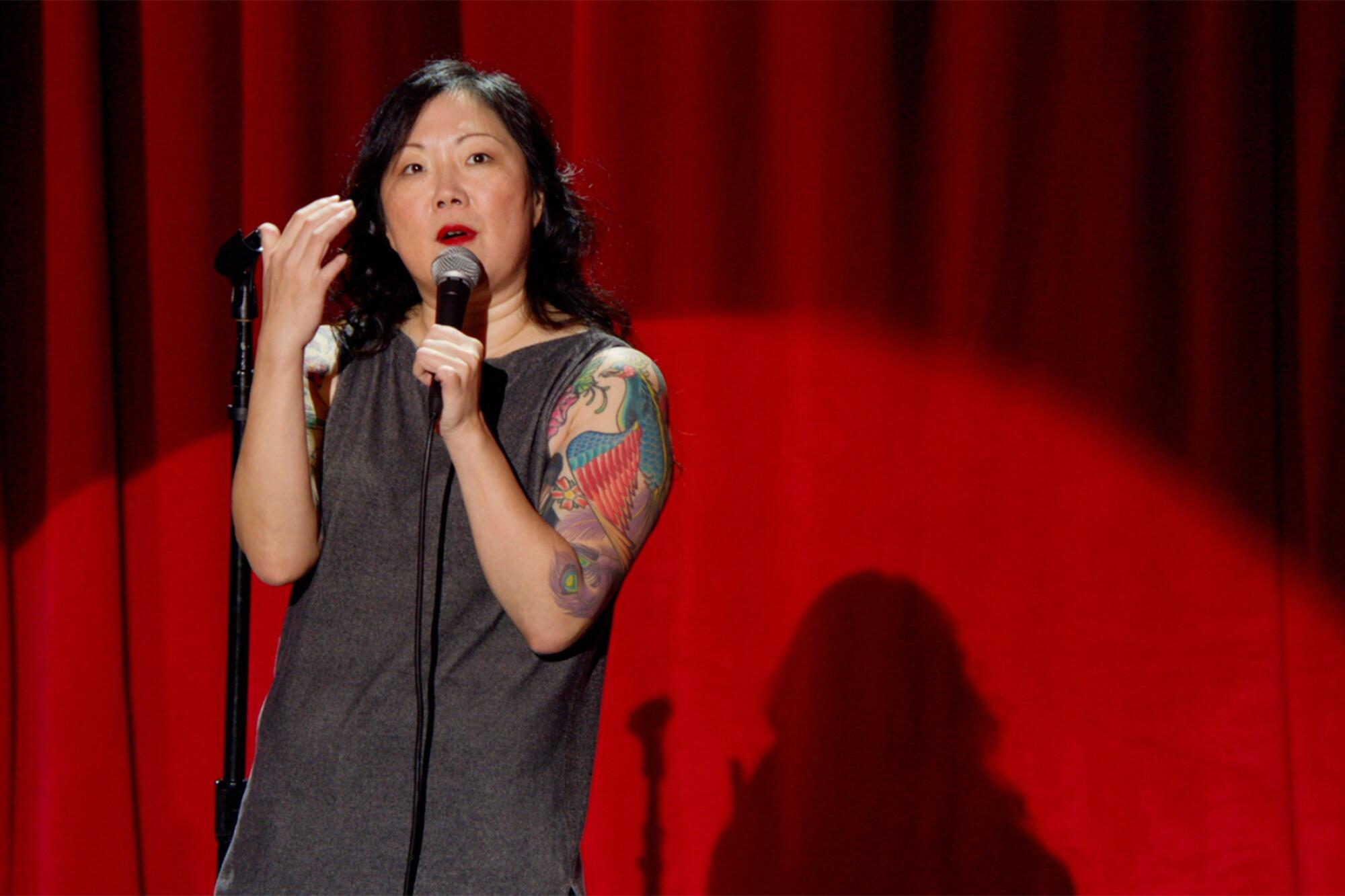Margaret Cho wearing black and speaking into a microphone in front of a red curtain