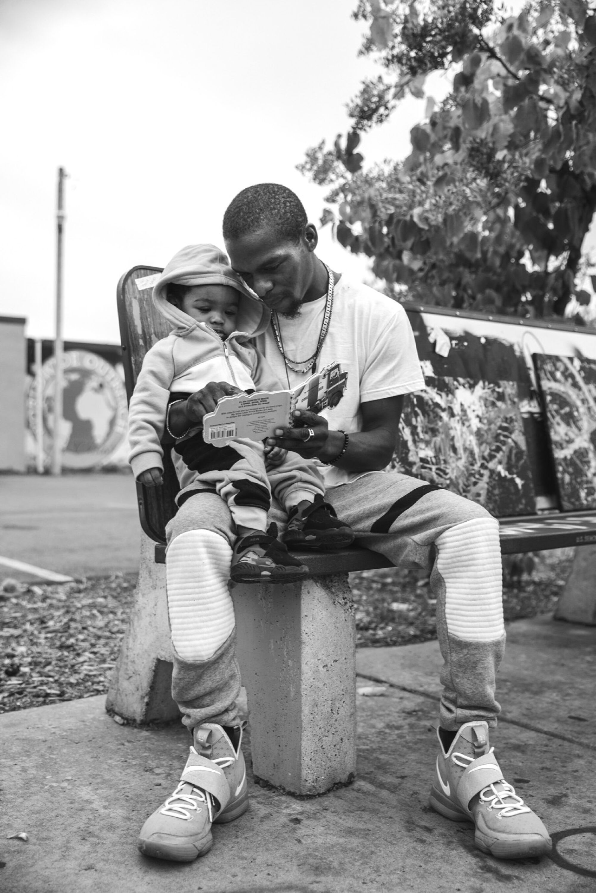 A man on a park bench reads a book to his son, who's sitting on his lap.
