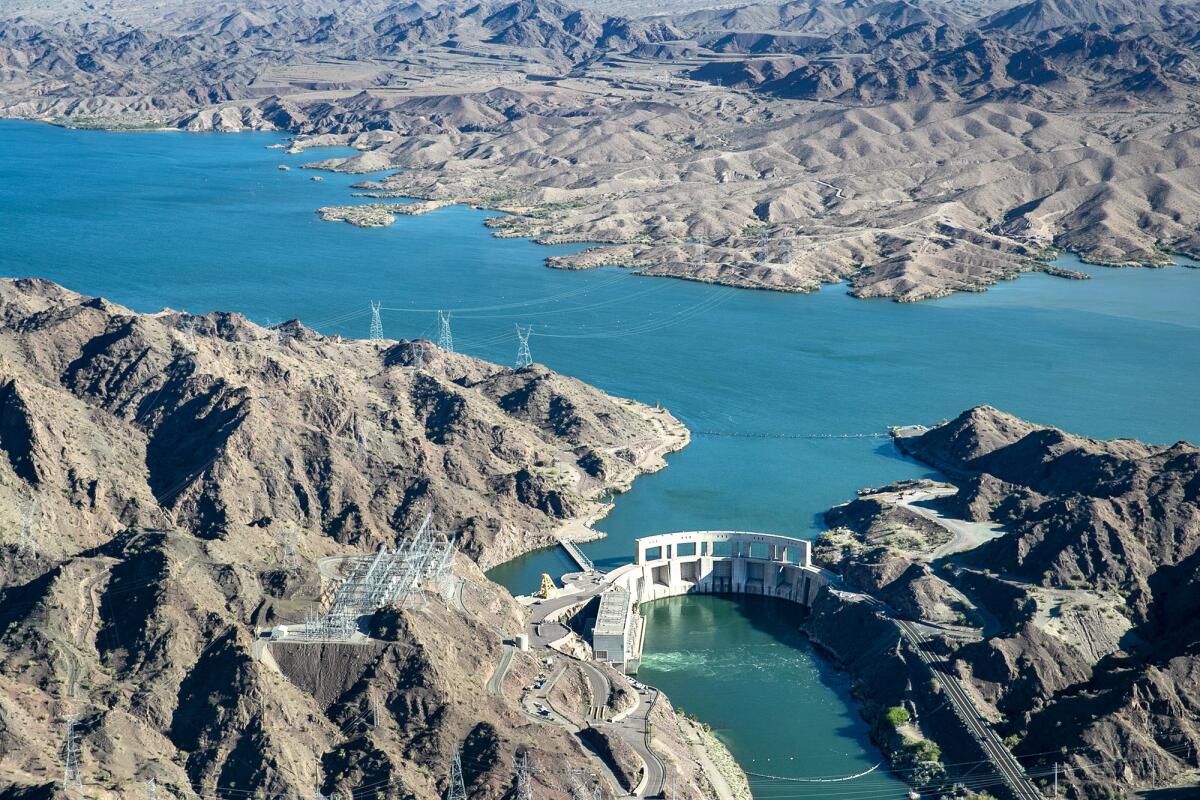 An aerial frame of a turquoise-water dam, filling a long stretch between two rocky, arid banks and landscape.