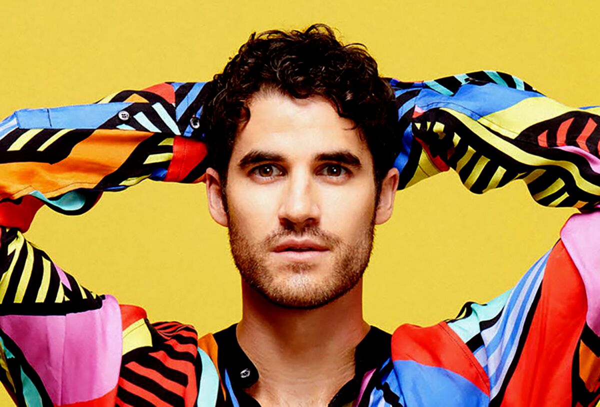 Darren Criss in a colorful pattern shirt with his hands behind his head in front of a yellow background.
