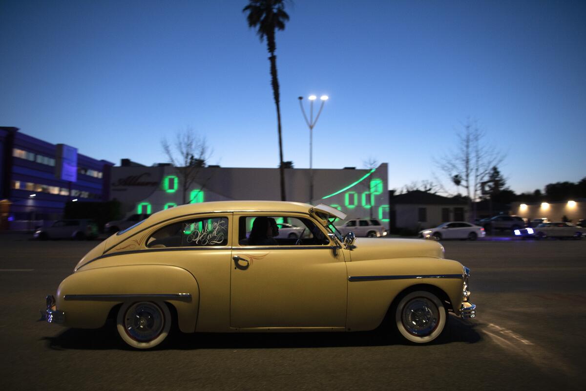 A 1940s style car with white walls cruises before a building illuminated with green neon at dusk