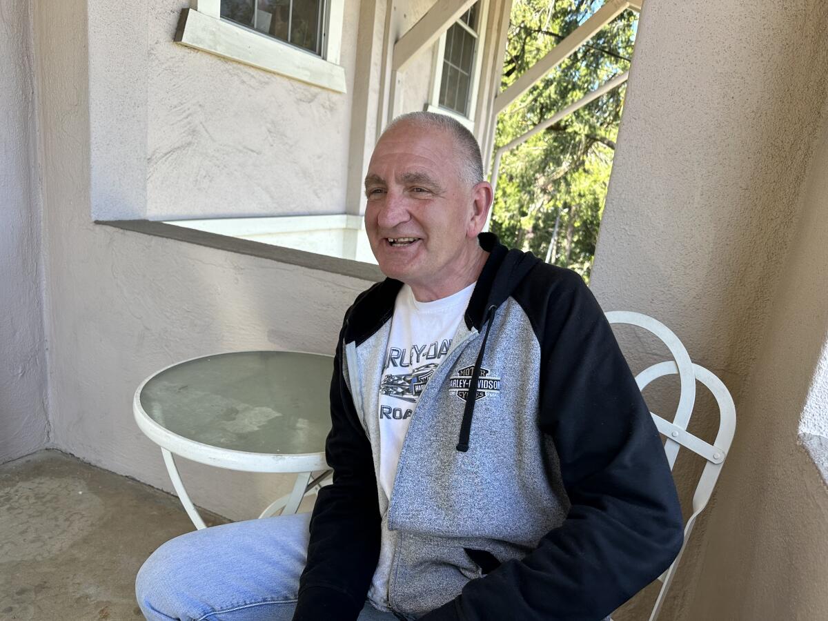 A balding man, seated and smiling, wears a sweatshirt and jeans.   