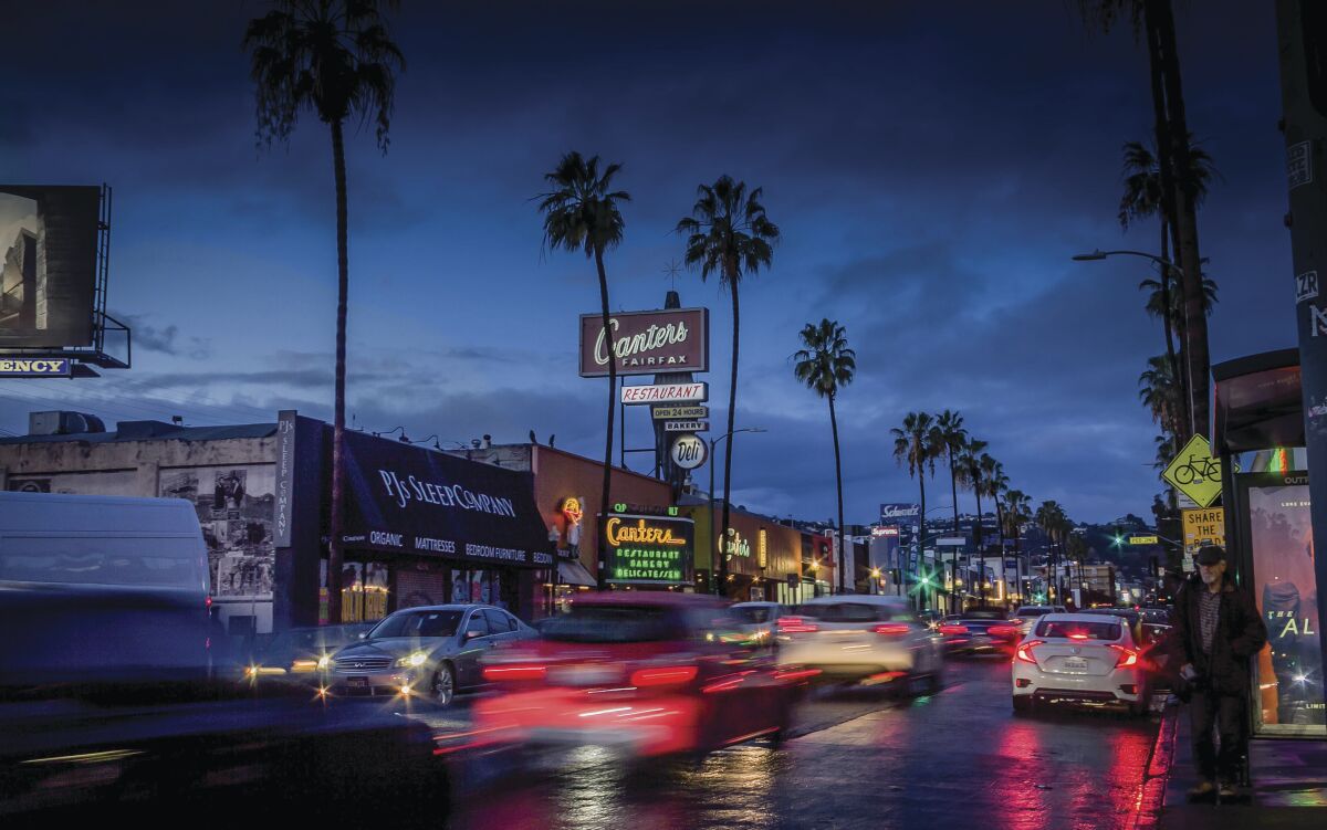 A sign that says "Canter's" is seen on a city street busy with traffic at dusk. Palm trees border the road.