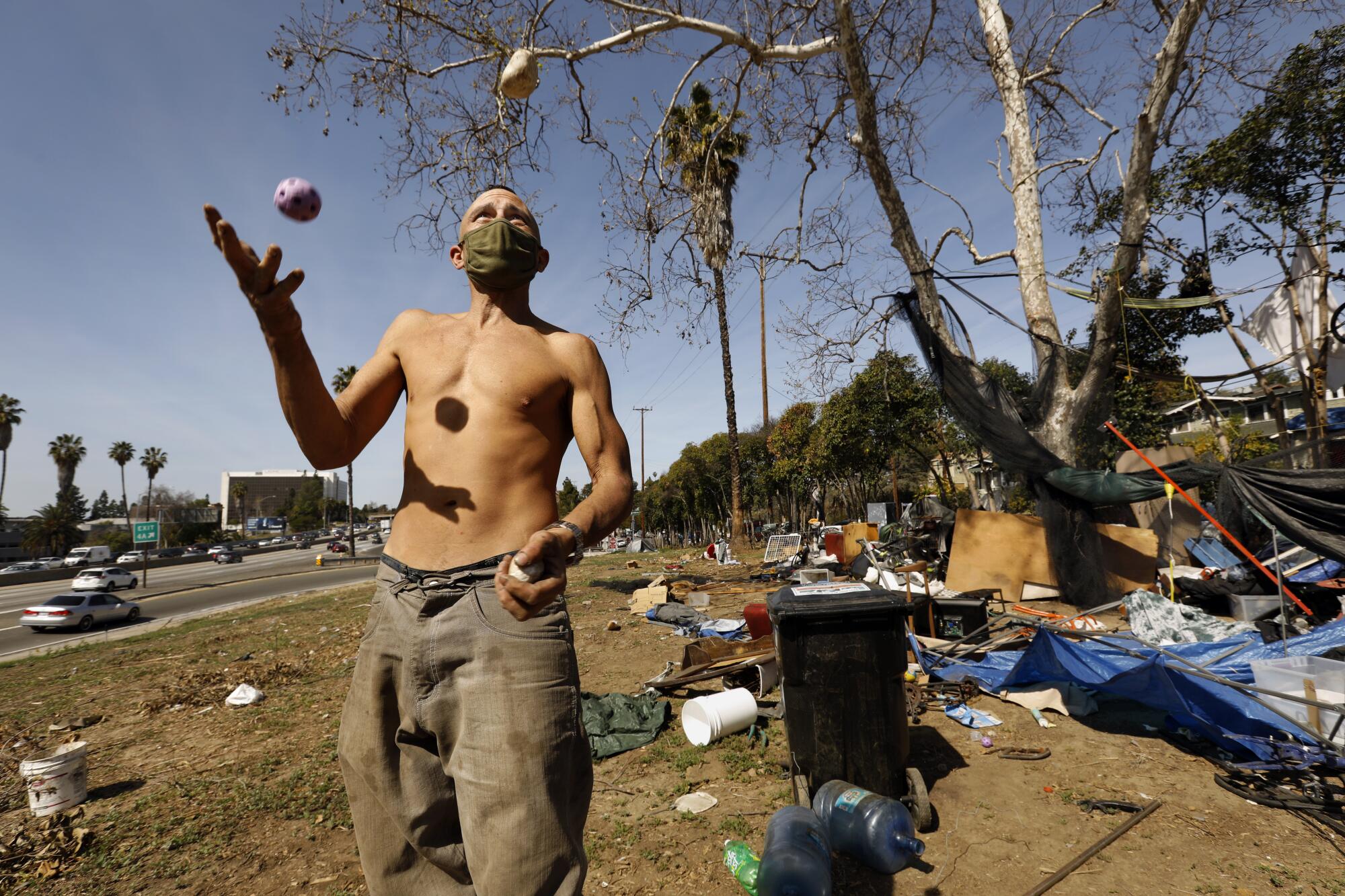 A shirtless man juggles near the freeway.