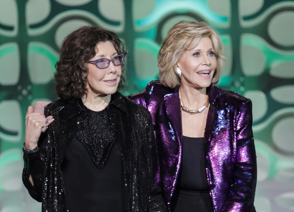 A mature woman stands on stage with her arm around the shoulders of another mature woman wearing glasses