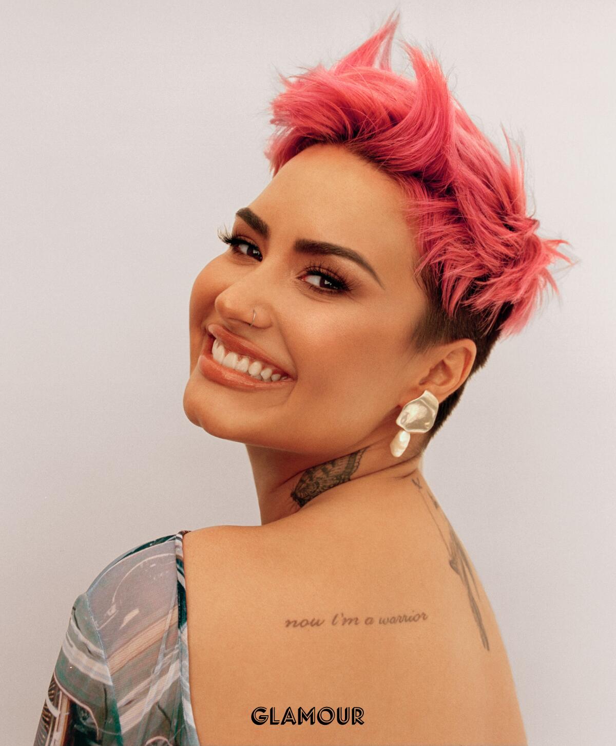 Demi Lovato, with short pink hair, looks over her shoulder at the camera