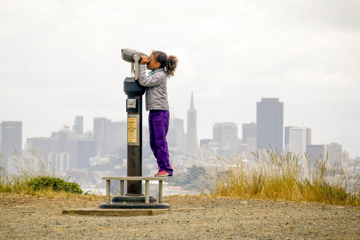 7-year-old Kayla LaMarche looks through a telescope with the San Francisco skyline in the distance behind her. 