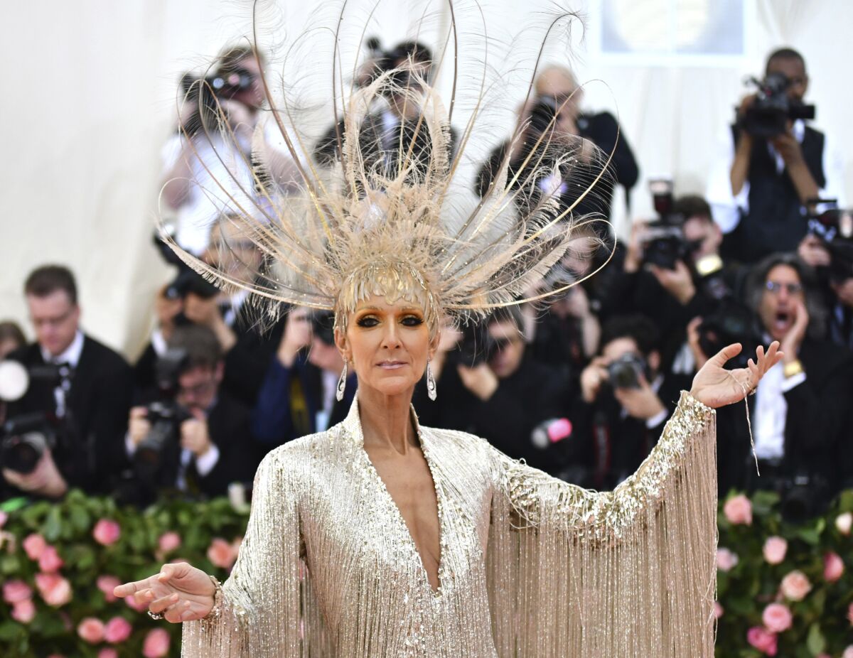 Celine Dion at wearing a gold gown with fringe sleeves, a large matching, feathered headpiece with her arms to the side