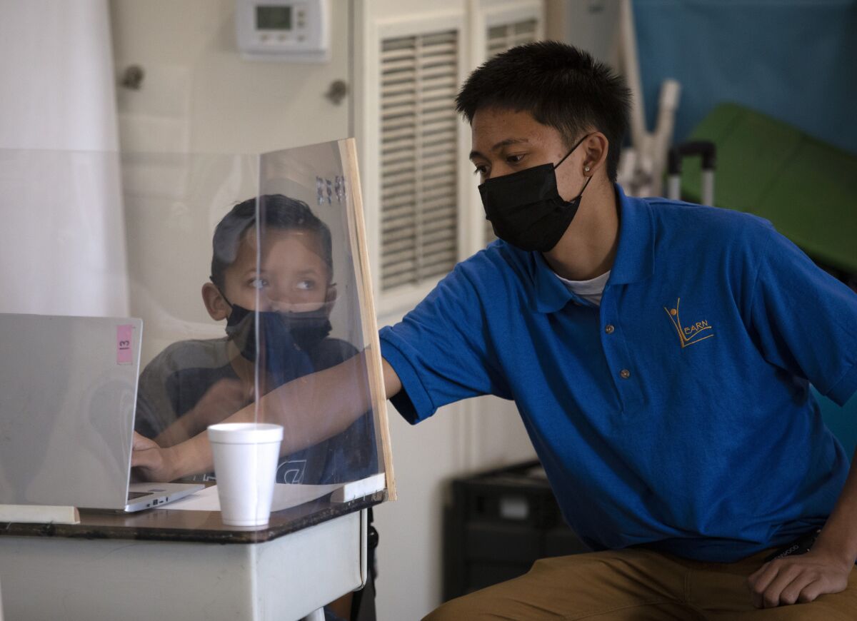 A school worker leans in to type on the laptop of a child sitting at a desk surrounded by acrylic plastic dividers.