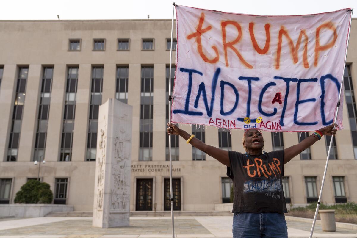 A person outside a courthouse holding a large sign and wearing a T-shirt that both read "Trump indicted again and again"