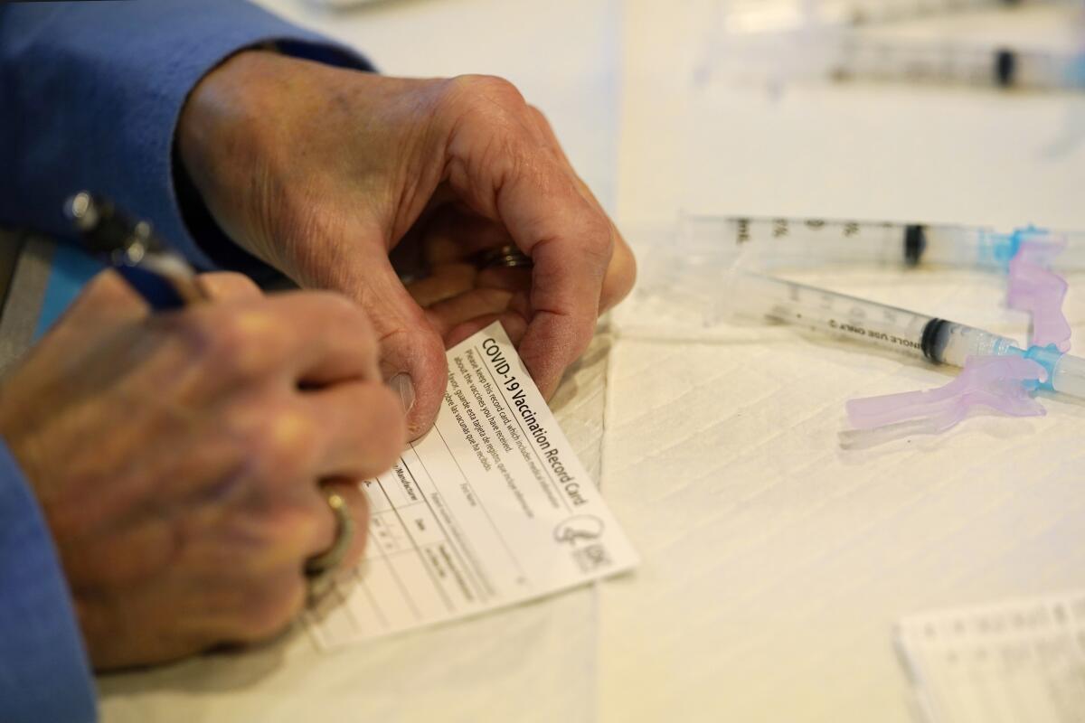 A closeup of someone writing with a pen on a card that says "COVID-19 Vaccination Record Card."