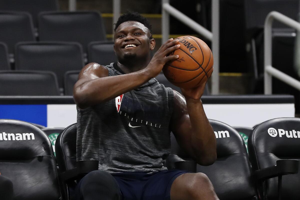 New Orleans Pelicans star Zion Williamson sits on the bench while holding up a ball and smiling.