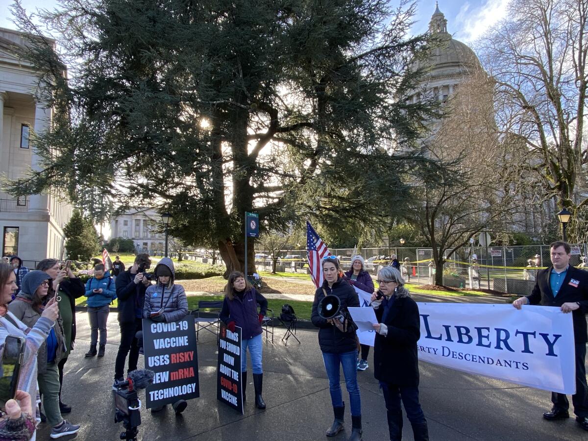 A small group of protesters stands in a public courtyard; a sign says "COVID-19 vaccine uses risky new mRNA technology."