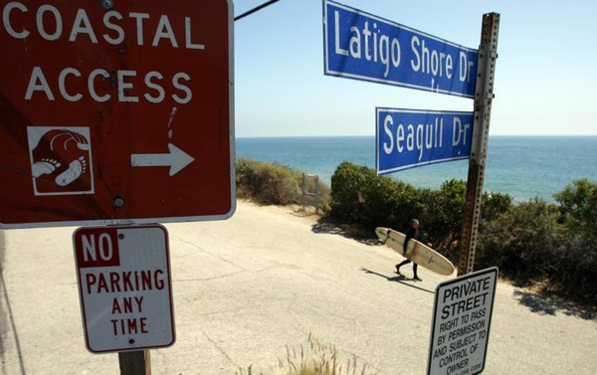 A surfer is framed by competing street signs regarding coastal access in Malibu. A bill pending before the state Legislature would give the Coastal Commission the authority to issue fines for access and other violations.