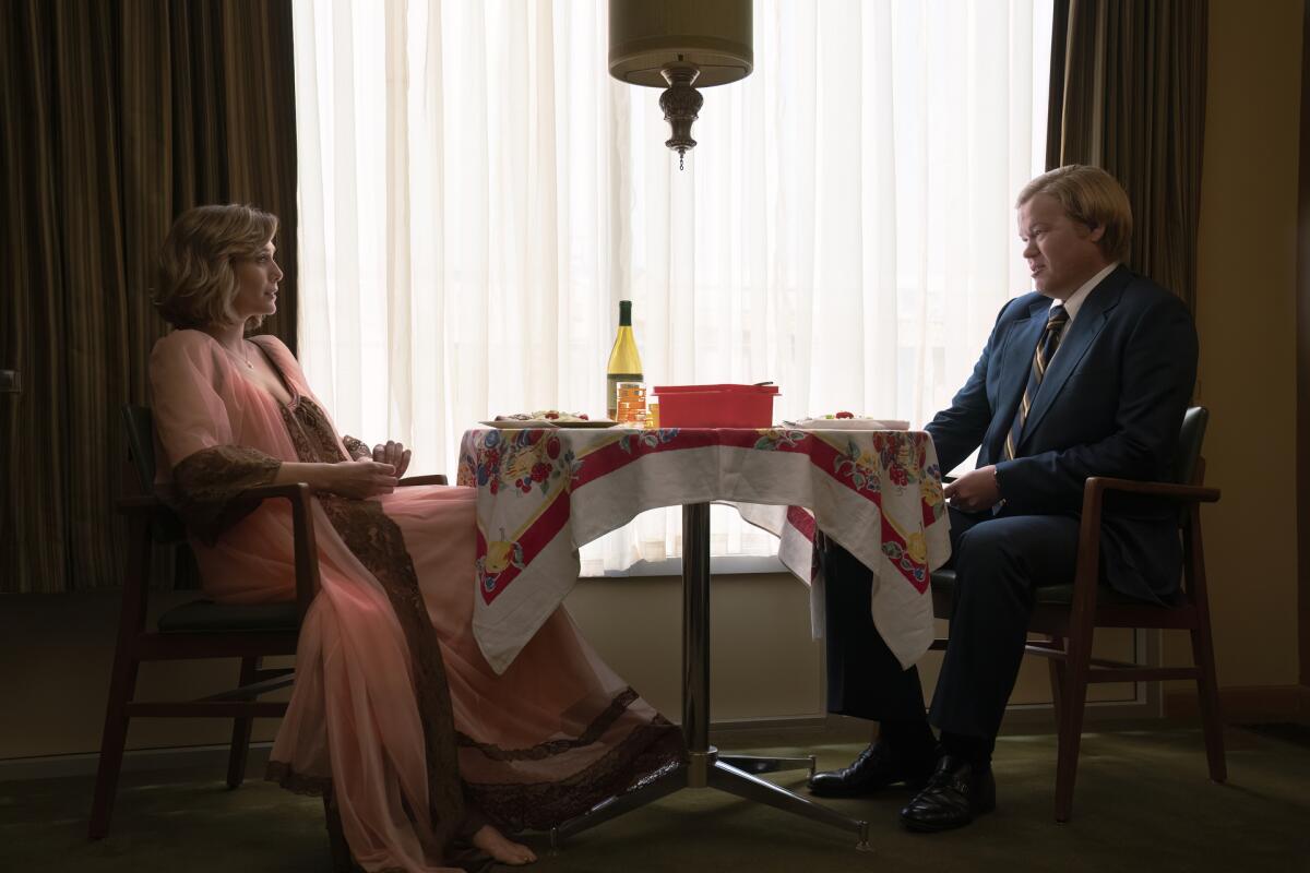 A woman and a man sitting across from each other at a small table near a large window with curtains