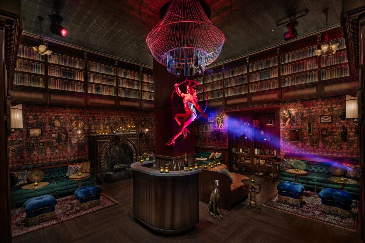 An aerialist in a spotlight hovers over a bar with benches and shelves of books.
