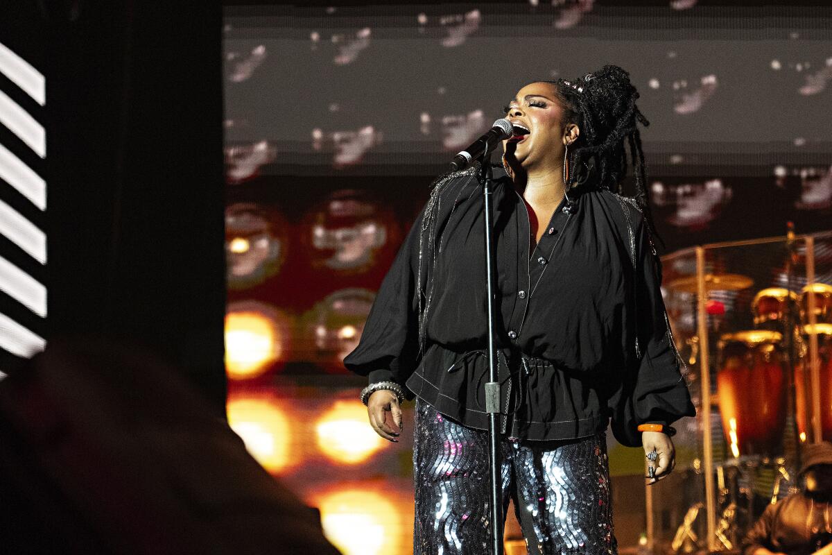 Jill Scott belting into a microphone on a stand while wearing a black button-down shirt and silver pants.