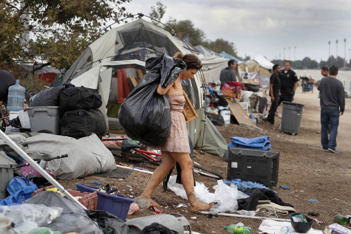 A woman caries large plastic bag and other items while walking barefoot in a homeless encampment.