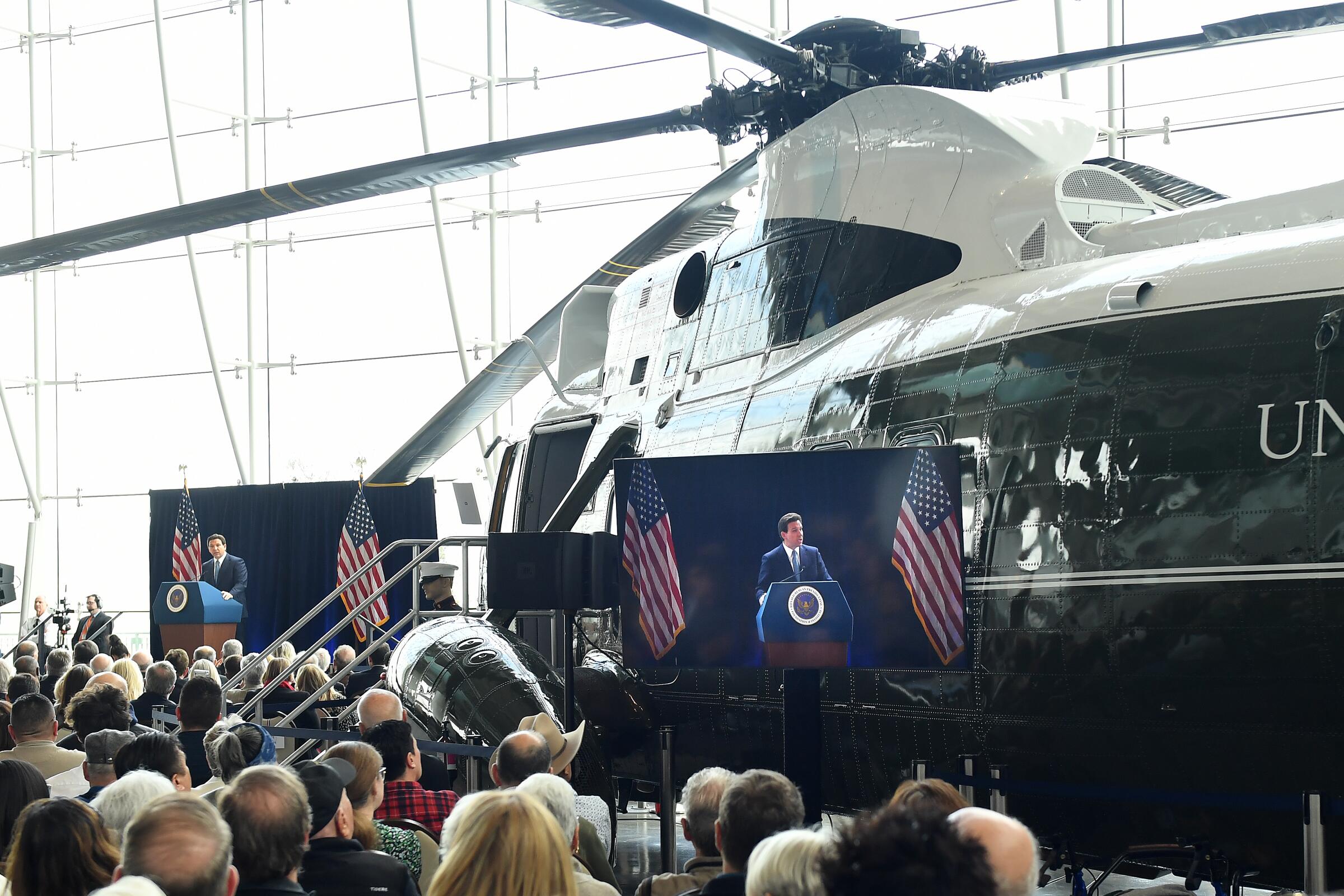 A man speaks from a podium to a crowd in front of a presidential helicopter in an exhibit hall