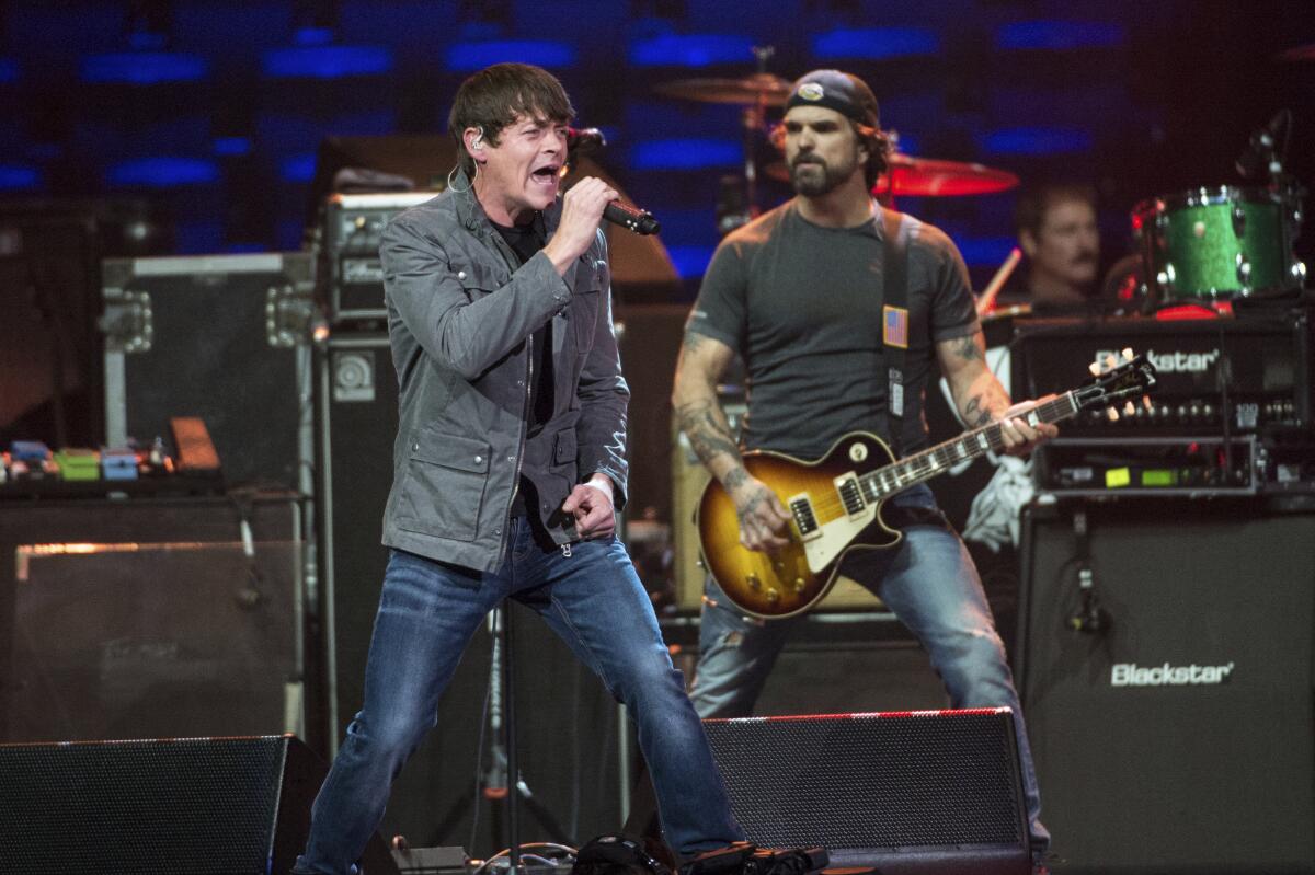 Two casually dressed rockers perform onstage with a band behind them.