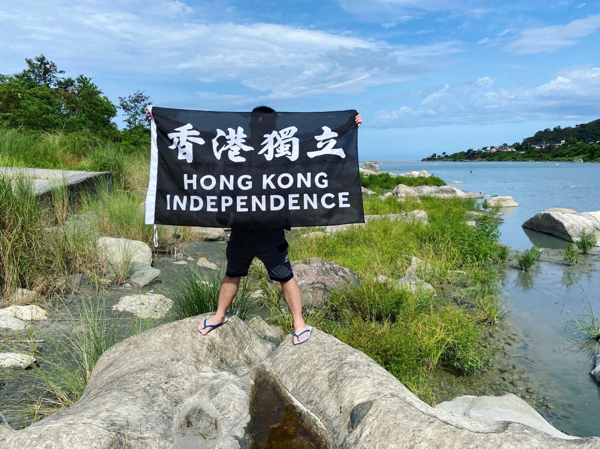 A young man standing on rocks near water holds a sign reading, "Hong Kong independence" 