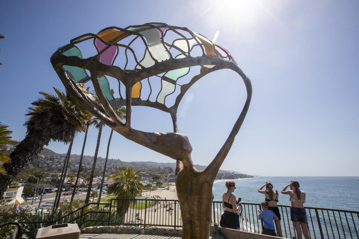 Behind a sculpture that incorporates colored glass, people stand at a railing with the ocean and beach below.