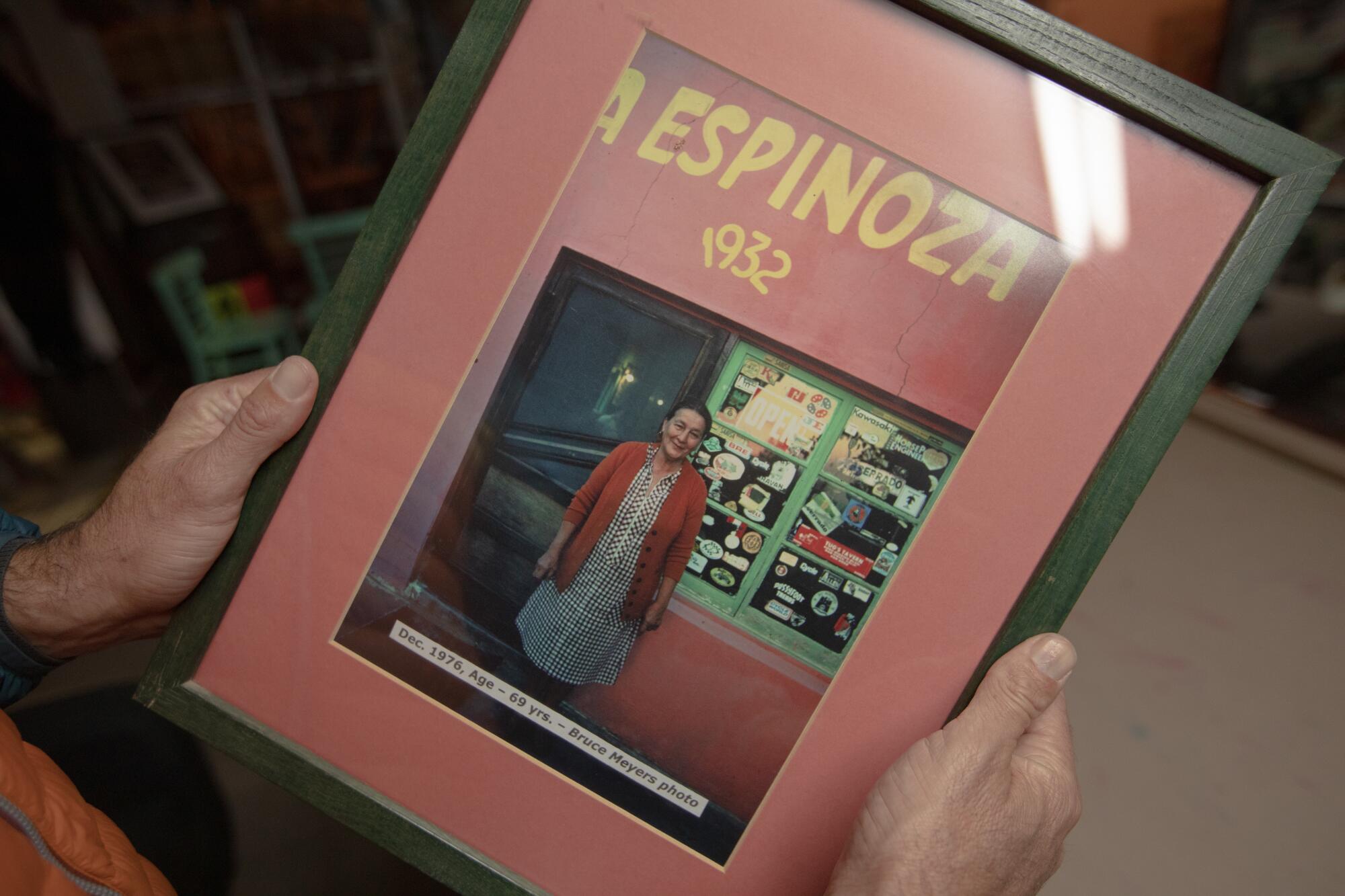 A pair of hands holding a frame photo of an older woman in front of a storefront, labeled "Mama Espinoza 1932".