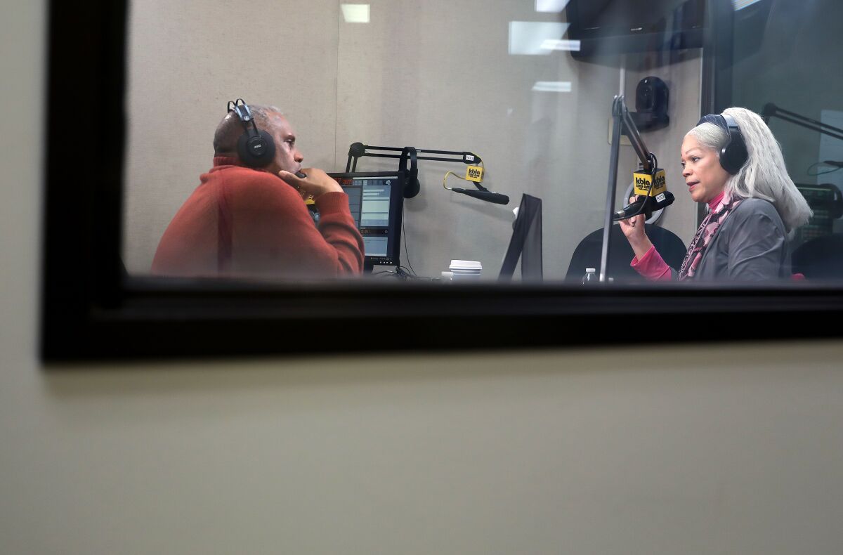 Two people talk into microphones in a room behind a window.