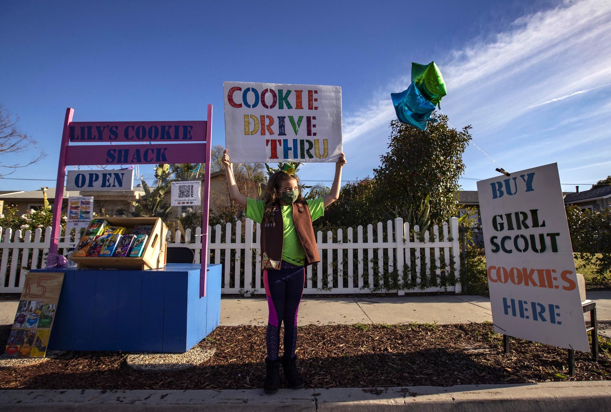 A girl in a Brownie vest holds up a sign that says "Cookie Drive Thru."