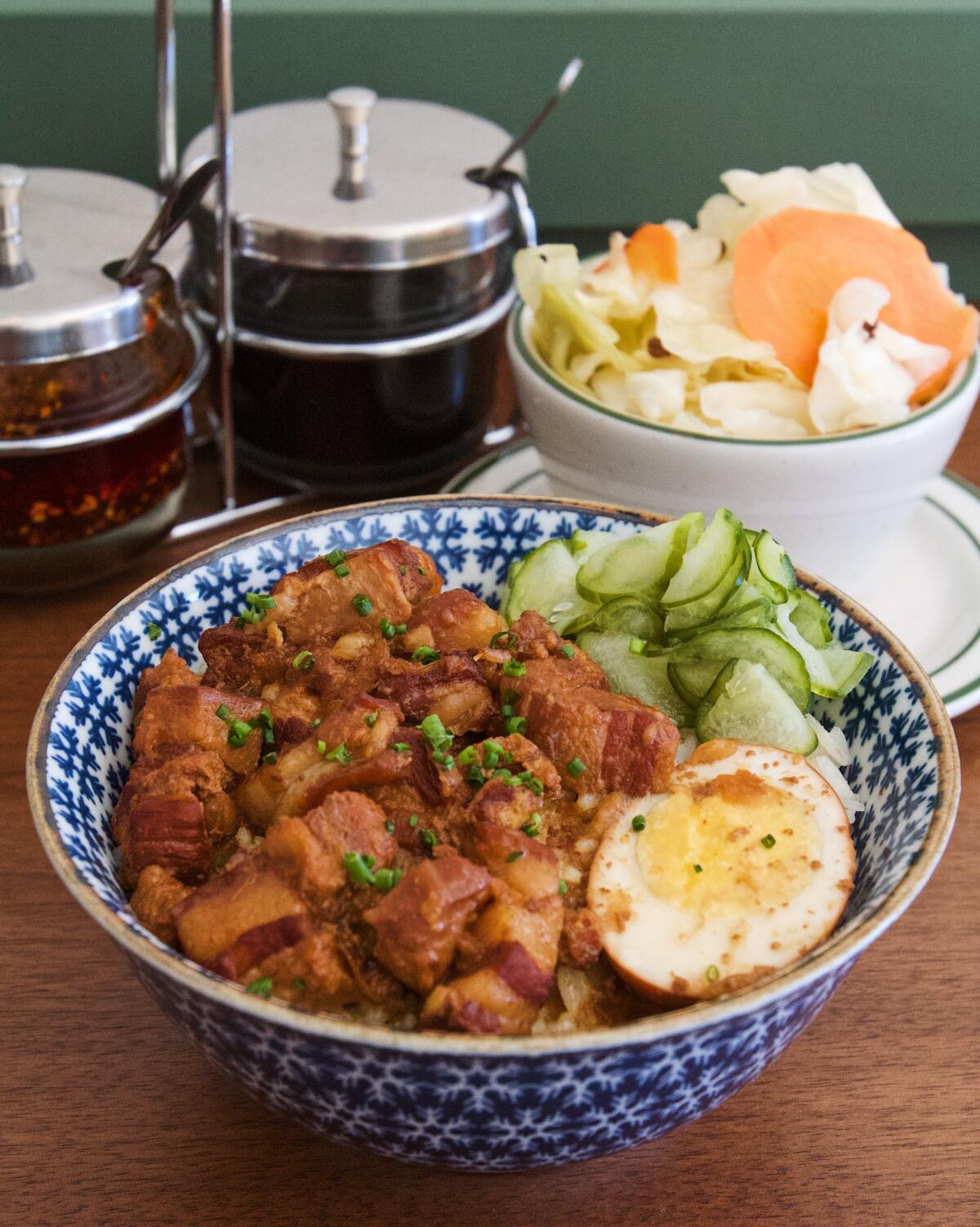 A vertical shot of a bowl of braised pork belly with rice and egg from Liu's Cafe. Condiments and pickled cabbage are behind.
