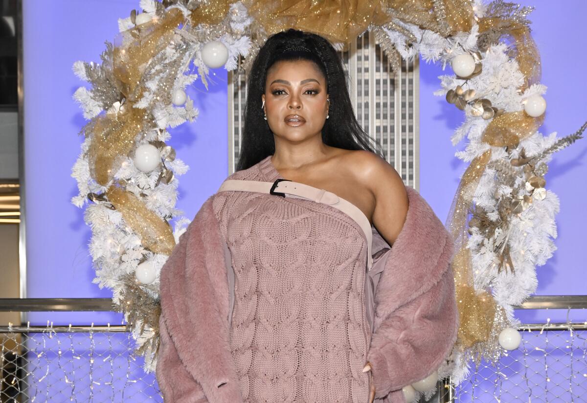 Taraji P. Henson, in lavender knit dress and fur jacket, poses in front of a purple backdrop and oversized wreath.