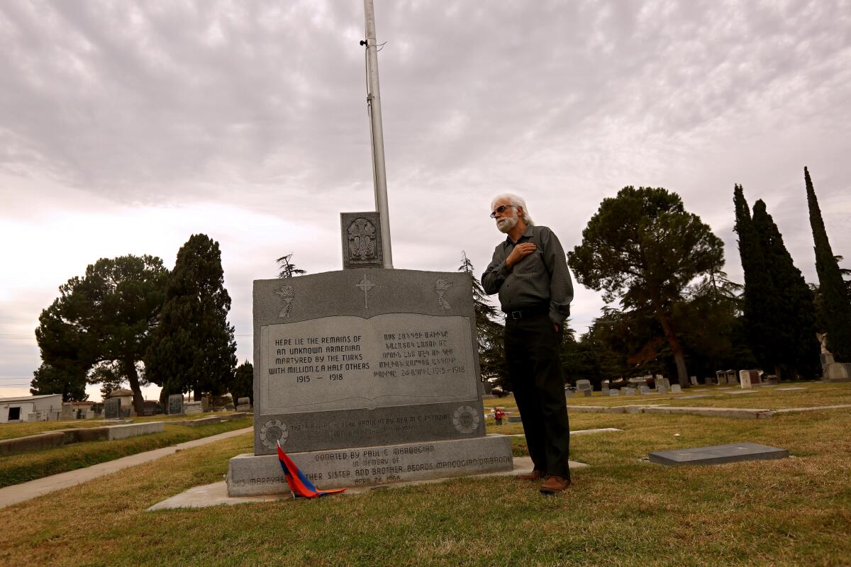 A man stands with his right hand over his heart next to a large headstone in a cemetery