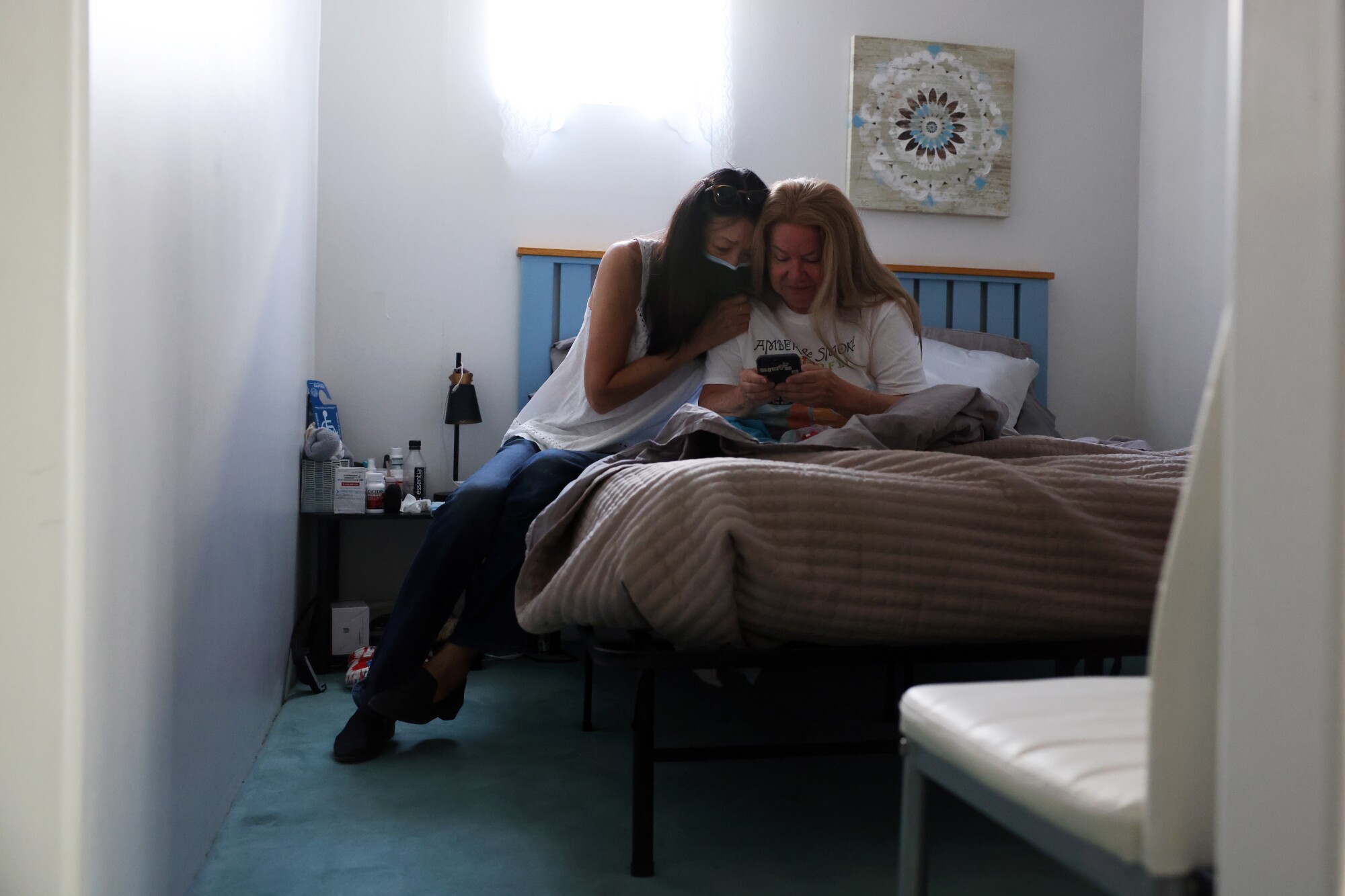 Two women sit on a bed and one holds a phone