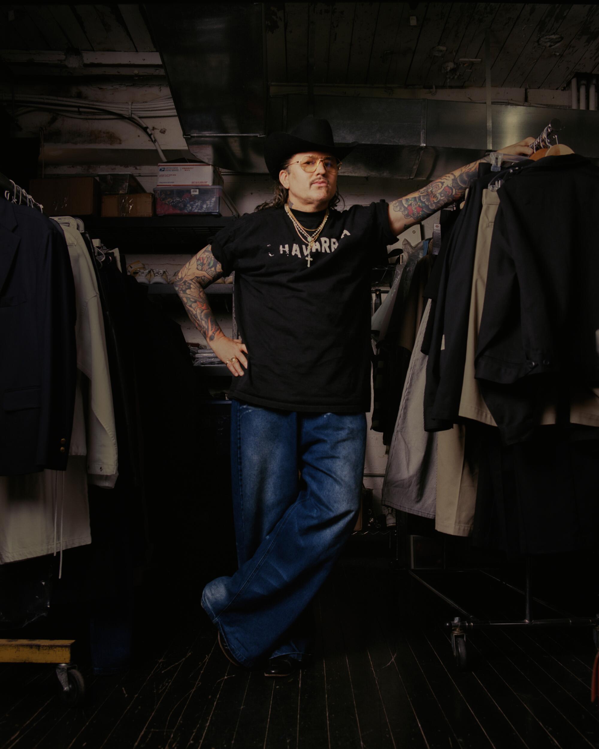 A man stands with his arm to his hip, leaning against a clothing rack.
