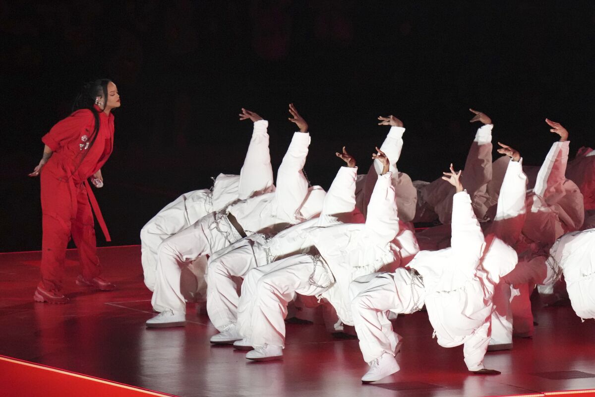 A woman in red stands in front of white-clad dancers bending backward, each with one hand in the air.