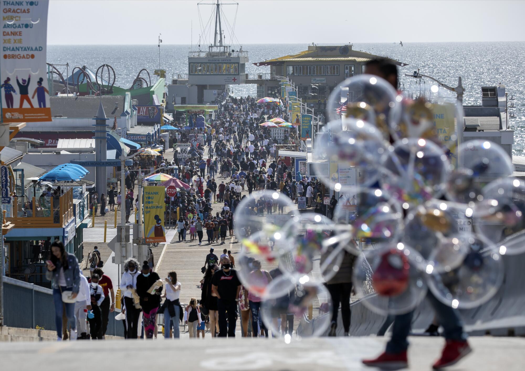 A balloon vendor, in the foreground on a pier crowded with people, is obscured by his balloons.