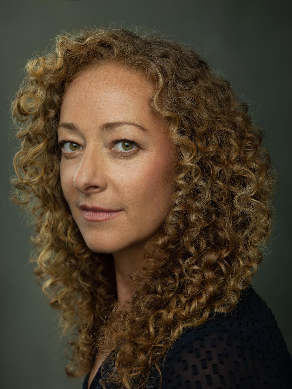 An author in profile with very curly dark blonde hair wearing black against a gray background.`