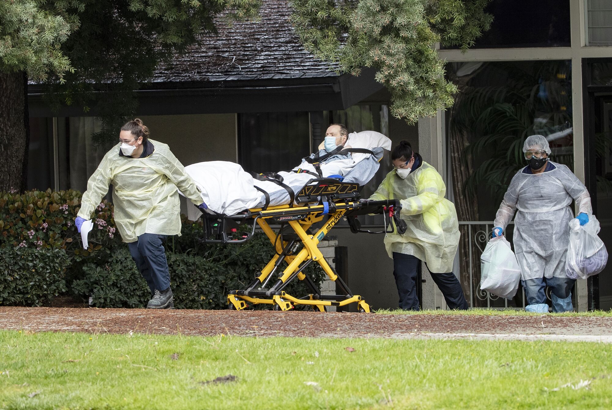 Two people in protective gear pull a stretcher with a patient on a sidewalk. Another carries two large plastic bags.
