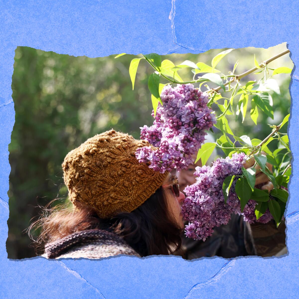 A woman in a hat and sunglasses leans in to smell purple flowers.