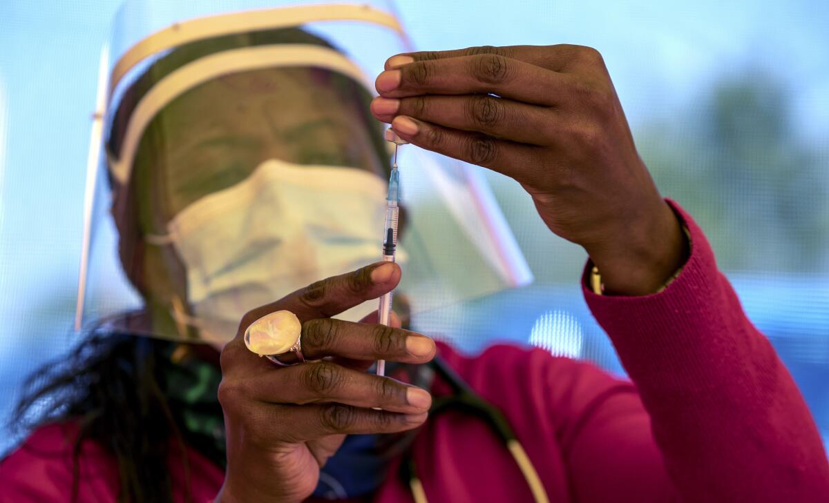 A woman in mask and face shield fills a syringe from a vial.