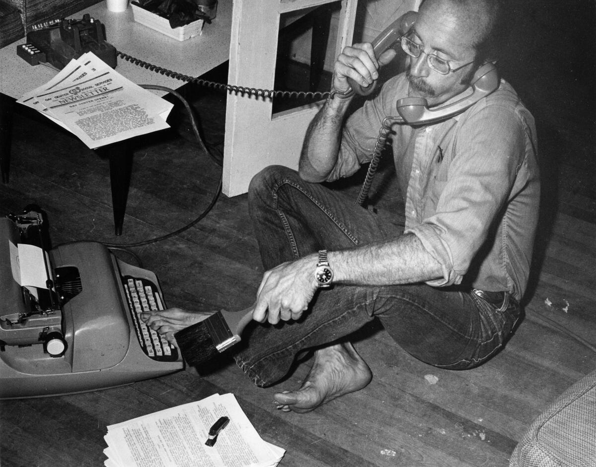 A man sits on a hardwood floor with legs crossed, holding a phone receiver in his right hand.