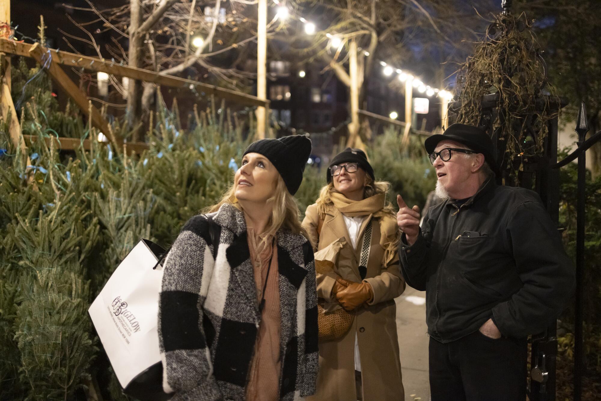 A man points while speaking to two women at a Christmas tree lot.