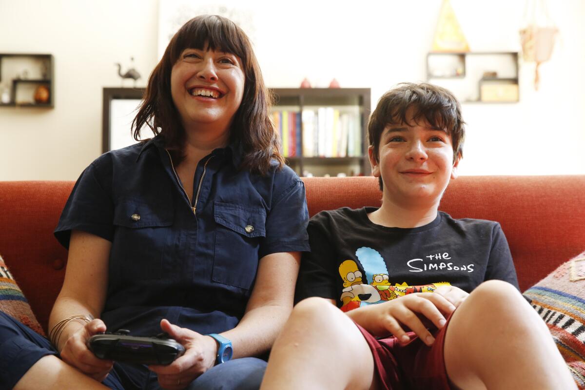 A woman holds a game console controller as she sits next to a boy on a couch.