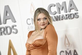 Elle King raises her left shoulder to her chin while posing in a tan dress at an awards show