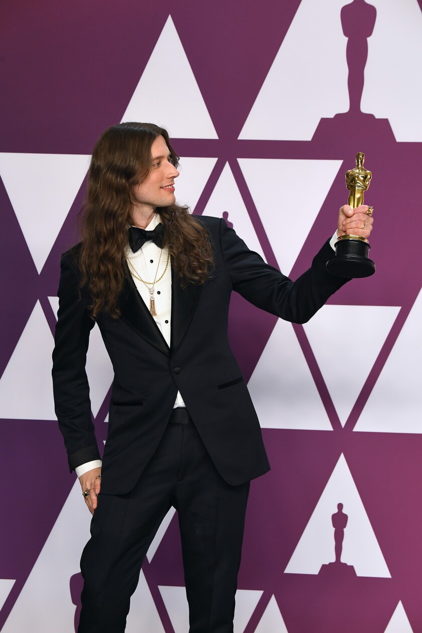 Composer Ludwig Göransson, in a navy blue tuxedo, holds up the Oscar he won for “Black Panther.”