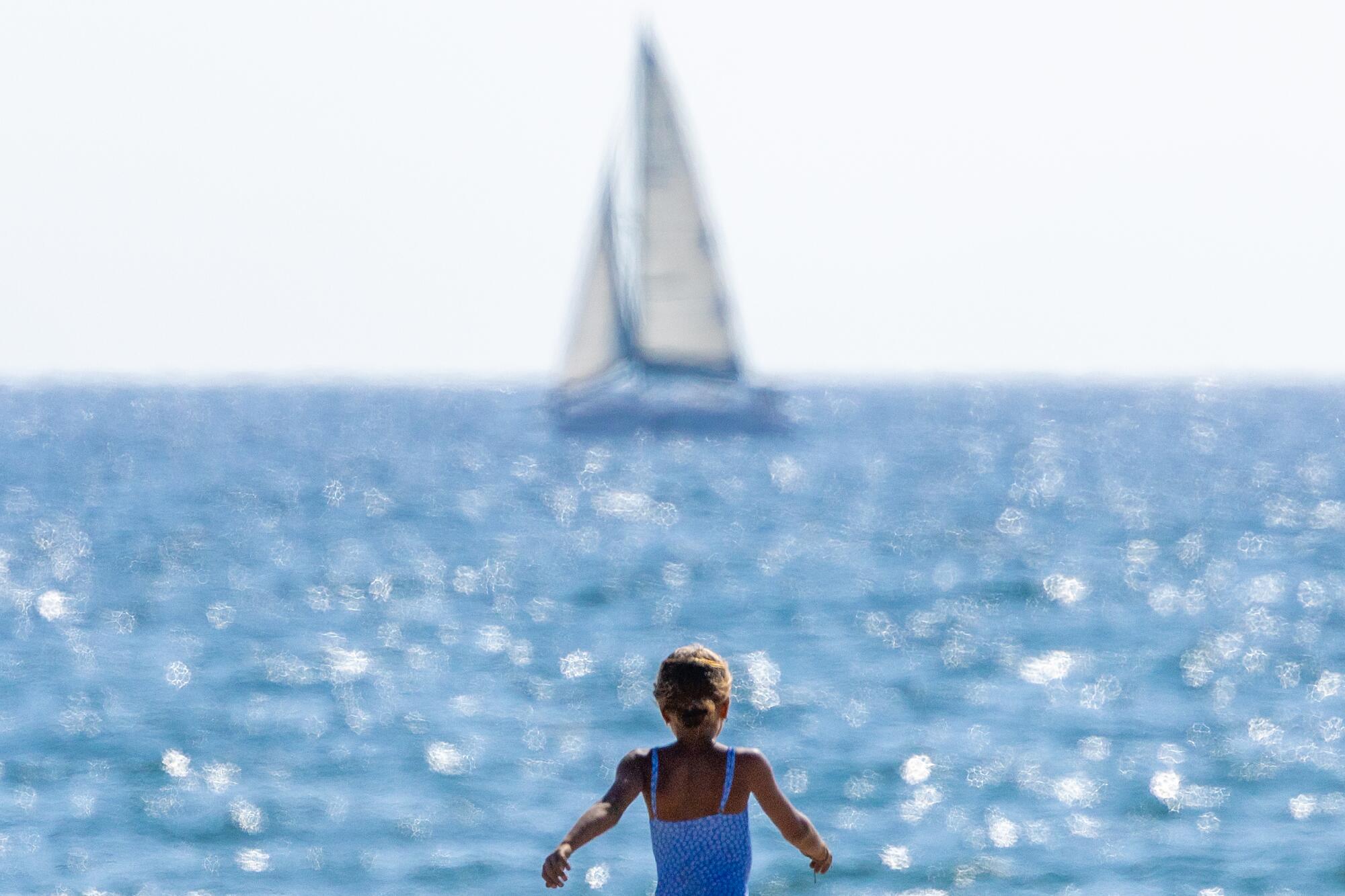 A beachgoer approaches the water as a sailboat floats in the distance.