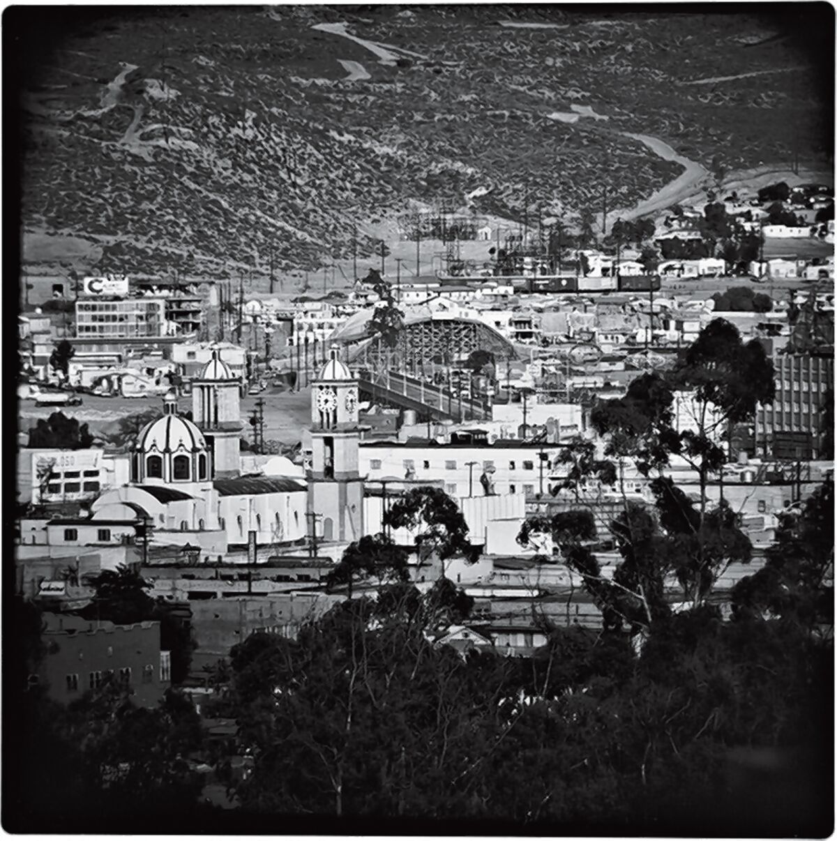 Guadalupe Cathedral in the foreground, with Puente Mexico and Puerta de Mexico (under construction) in the center of the photograph. In the background, the hillside with vegetation in San Ysidro, California, and to the right a portion of Colonic Libertad. Gelatin Silver Print 1964. Photograph by Harry Crosby, Harry Crosby Collection, Special Collections & Archives, UC San Diego